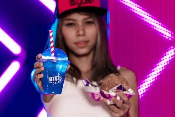 foto de menina desfocada com copo transparente em primeiro plano com liquido azul e segurando hamburguer na outra mão