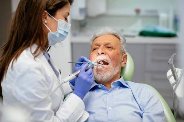 Dentista consulta idoso deitado em cadeira num consultório odontológico, examinando sua boca com ferramentas. Ela usa máscara e jaleco - Metrópoles