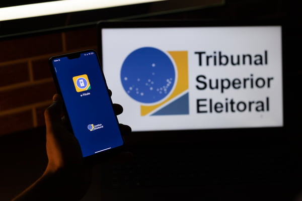 mão segura celular aberto no aplicativo do e-titulo desfocado e ao fundo a logo do tribunal superior eleitoral.