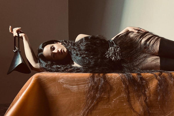 A cantora Rosalía, uma mulhe jovem, branca, de cabelo ondulado longo, posando para a campanha da marca Acne Studios. Ela está deitada em uma mesa e usa um casaco preto de couro, um cinto com fivela prata e uma bolsa de couro preto.