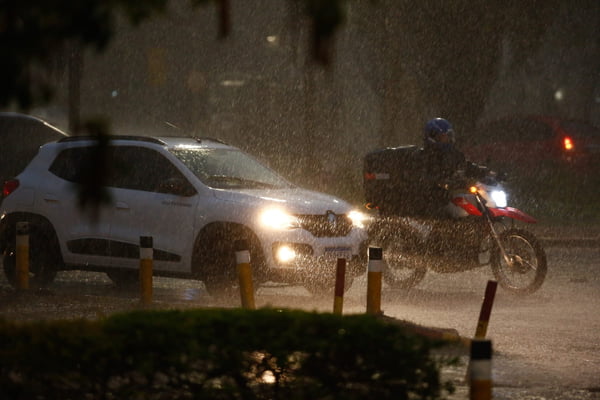 Carro branco e motociclista em moto vermelha passam por rua de Brasília, em tarde chuvosa. Farol dos dois veículos iluminam a rua, com calçada e arbusto desfocados e em primeiro plano - Metrópoles