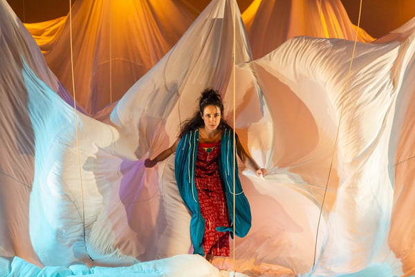Mulher com vestido vermelho e manta azul com um pano claro ao fundo na montagem cênica À Beira do Sol no Teatro Dulcina – Metrópoles