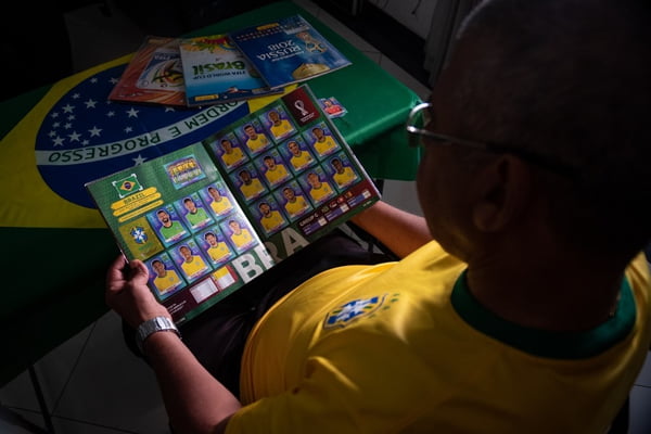 Homem com camisa do Brasil sentado, olhando para baixo, onde segura um álbum de figurinhas da Copa do Mundo nas mãos