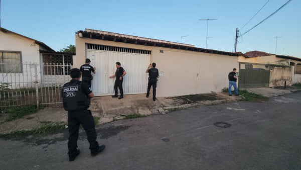 Quatro policiais civis do Distrito Federal, vestidos com uniformes pretos, em rua, em frente a casa de portão branco e muro bege. O imóvel fica entre duas outras casas