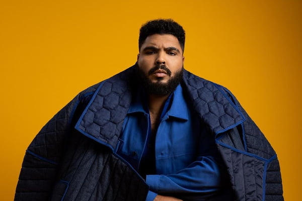 O humorista Paulo Vieira posa para foto com jaqueta estilizada, de braços cruzados sob fundo amarelo - Metrópoles