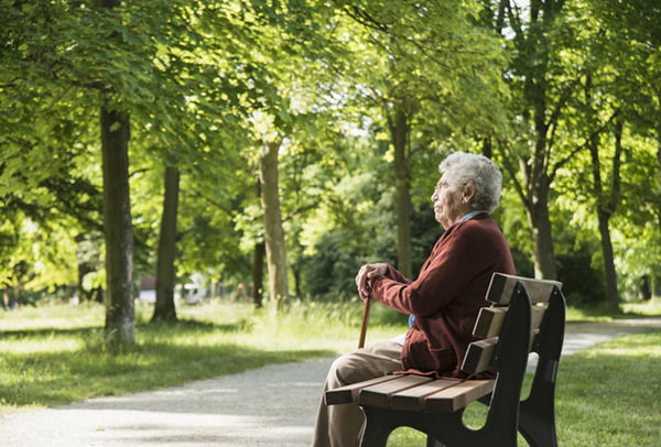 Imagem colorida: idosa solitória sentada em banco de parque com uma bengala - Metrópoles