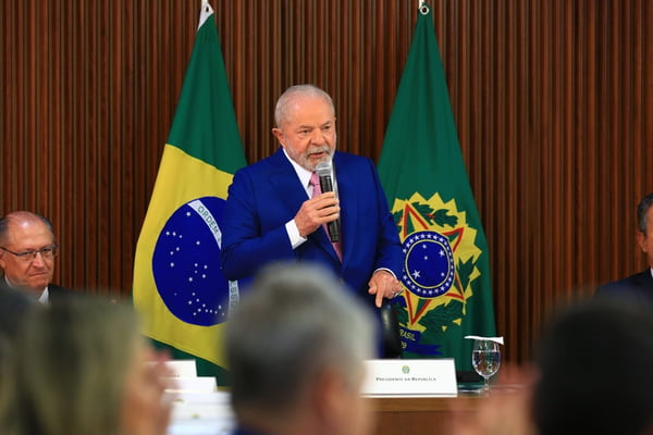 O presidente Lula faz primeira reunião ministerial no Palácio do Planalto e pede boa relação com o Congresso Nacional. Na imagem ele está de pé, na ponta de uma mesa, falando em microfone com bandeiras do Brasil atrás e ao lado de Alckmin - Metrópoles