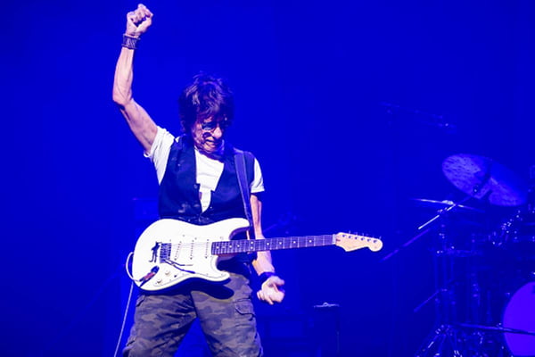 Jeff Beck durante show comguitarrada pendurada ao pesço enquanto levanta o punho direito e cabeça baixa
