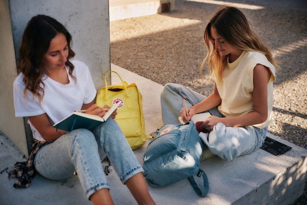 Duas adolescentes brancas, com cabelo liso, sentadas no chão estudando. Elas usam calças jeans e blusas beges e escrevem em cadernos. - Metrópoles