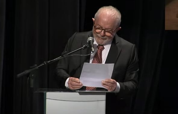 O presidente Lula discursa em púlpito, lendo folha de papel, sobre países devedores do BNDES durante cerimônia - Metrópoles