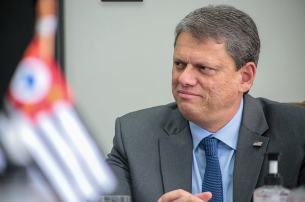 O governador de São Paulo, Tarcísio de Freitas, durante reunião em seu escritório. Ele sorri, olhando para o lado, com a bandeira do estado desfocada a frente - Metrópoles
