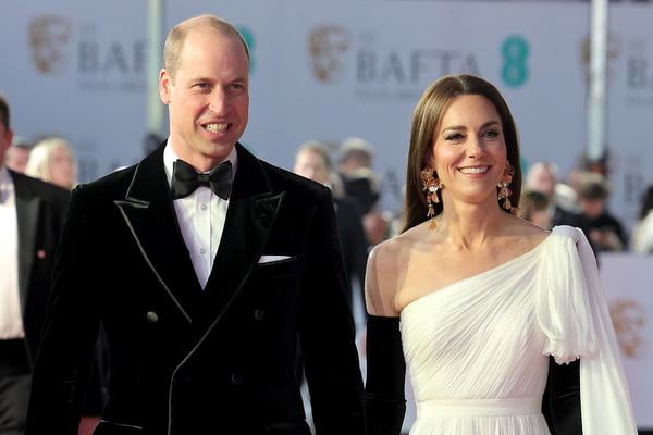 Princesa Kate Middleton usa cabelo partido ao meio e brinco maxi de flores douradas da Zar. Ela usa um longo vestido branco e luvas pretas. Ao lado está o príncipe William com um smoking preto e gravata borboleta. Ele é um homem branco e careca- Metrópoles