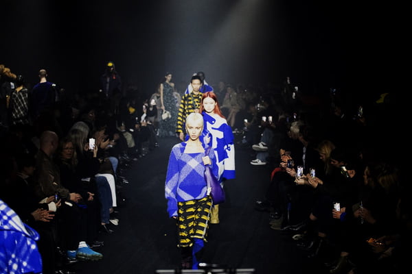 Em desfile, fila de modelos em meio à plateia. A modelo da frente usa look estampado em xadrez com blusa e suéter em azul e branco, além de saia em amarelo e preto - Metrópoles