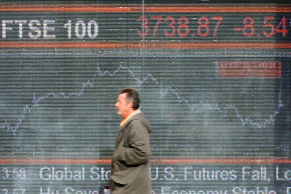Imagem de painel da Bolsa de Valores de Londres, exibindo números do índice FTSE 100 e, em frente, um homem de terno escuro caminhando - Metrópoles