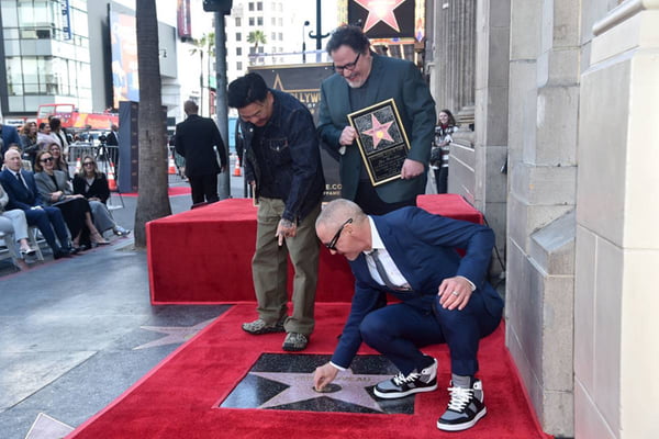 Robert Downey Jr. agachado colando chiclete em estrela da calçada da fama enquanto dois homens observam de pé - metrópoles