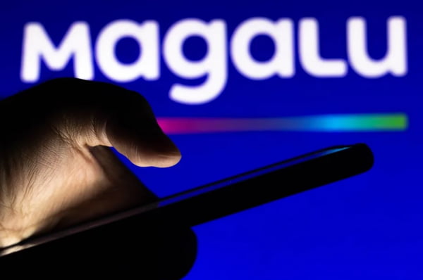 Imagem colorida mostra a silhueta de uma mão segurando um celular e o logotipo do Magazine Luiza, escrito Magalu, ao fundo - Metrópoles