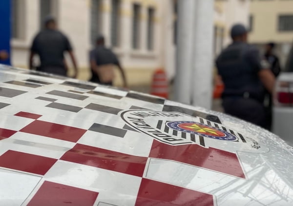 Imagem colorida do capô de uma viatura da Polícia Militar de SP. A peça é quadriculada em branco e vermelho com o logo da PM paulista. Ao fundo, alguns PMs - Metrópoles