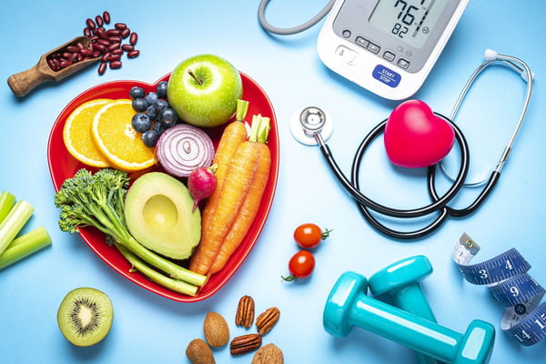Foto colorida de superfície azul com estetoscópio, frutas dentro de um prato em formato de coração, equipamento para medir pressão arterial, peso para fazer exercícios e fita métrica - Metrópoles