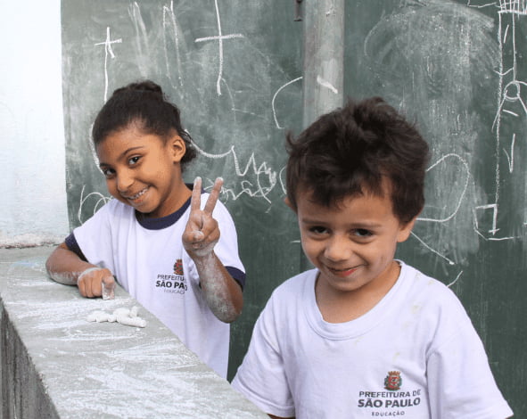 Duas crianças com uniforme escolar da rede municipal de SP brincam com giz de cera e acenam para a foto - Metrópoles