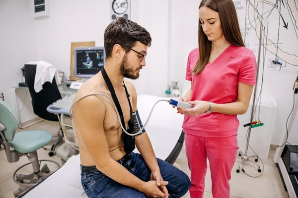 Foto mostra enfermeira vestida de rosa medindo a pressão cardíaca de um homem sem camisa - Metrópoles