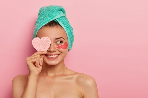 Mulher com toalha na cabeça fazendo auto cuidado, usando bolsa de olheiras nos olhos e segurando um coração com a mão - Metrópoles