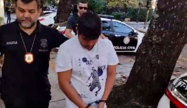 Torcedor do Flamengo de camiseta branca e cabeça baixa é conduzido por dois policiais até uma delegacia do Rio de Janeiro