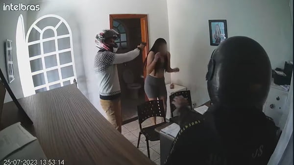 Homem com capacete de moto aponta arma para mulher dentro de uma sala. Outro homem com capacete de moto e outro homem com casaco cinza estão na sala