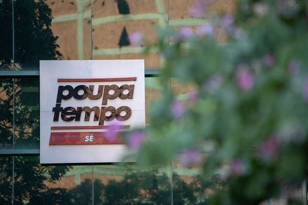Imagem colorida da fachada do Poupatempo Sé. A placa com o nome do local está atrás de uma árvore com folhas verdes e folhas rosas desfocadas - Metrópoles