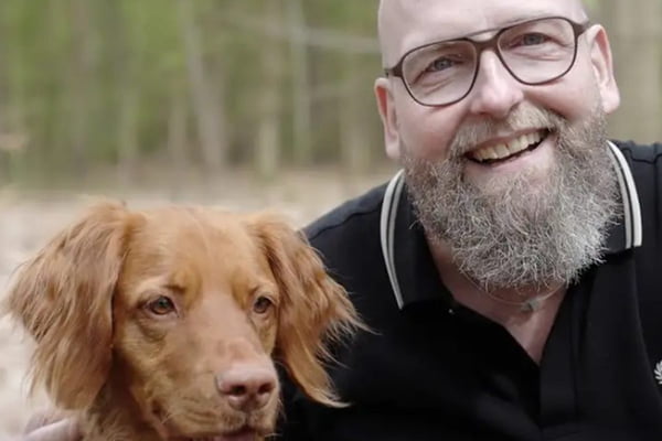 Imagem colorida mostra homem careca de barba e óculos sorrindo e cachorro marrom - Metrópoles