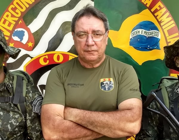 Homem com camiseta marrom, cabelos grisalhos e óculos, de braços cruzados - Metrópoles