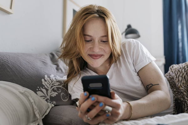 Foto colorida de uma mulher deitada no sofá com o celular na mão e sorrindo olhando para a tela dele - Metrópoles