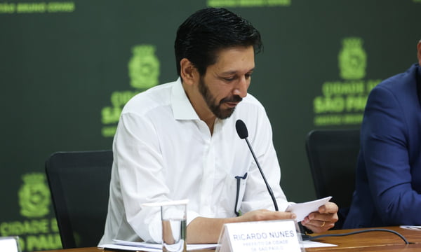 imagem colorida mostra homem branco, com barba e cabelo pretos. ele veste camisa branca, está sentado e lê um papel em frente a um microfone. metrópoles