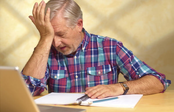 Foto colorida de homem idoso sentado em frente a computador e caderno com mão na testa - Metrópoles