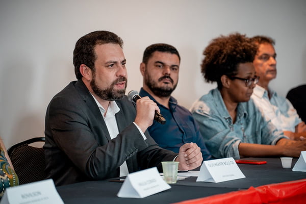 Imagem colorida mostra Guilherme Boulos falando ao microfone, sentado, em uma mesa de reunião com aliados - Metrópoles