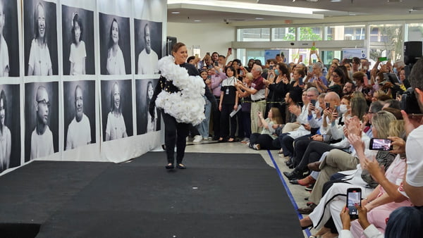 Imagem colorida mostra mulher desfilando em passarela montada em hospital. Ela veste preto e tem uma roupa feita de papel branco por cima do traje. dezenas de pessoas acompanham o desfile e a aplaudem - metrópoles