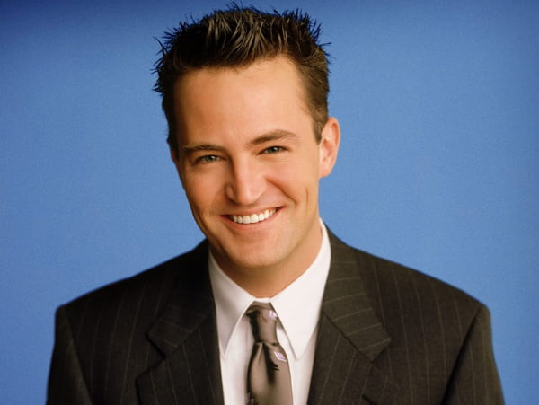 Foto colorida de Matthew Perry vestido de terno com fundo azul - Metrópoles