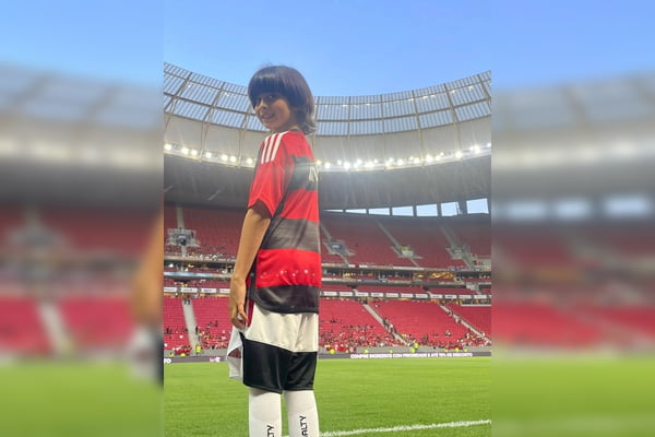 Foto colorida de menino em estádio; ele é branco, tem cabelos castanhos, veste uniforme de futebol do Flamengo e olha para a câmera