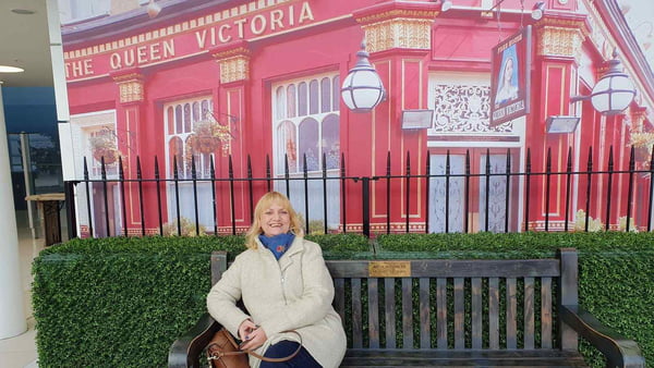 Foto mostra Lesley-Ann Woodhams sentada em um banco público com uma paisagem inglesa ao fundo. Ela preveniu o câncer de mama tomando o remédio anastrasol