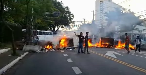 imagem colorida mostra duas pessoas em frente à barreira montada em avenida de são paulo. há fogo nos objetos espalhados pela via - metrópoles