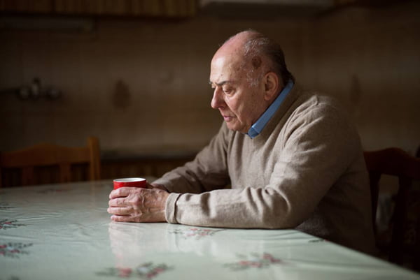 Imagem colorida de idoso sentado sozinho em mesa enquanto segura xícara - Metrópoles