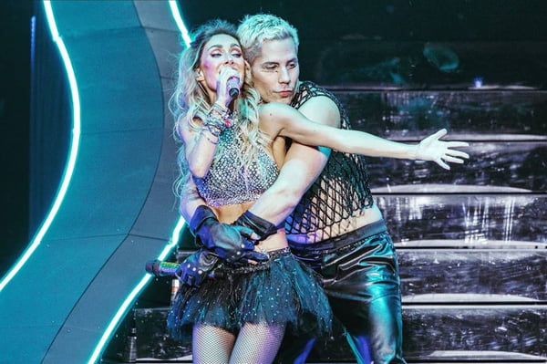 Foto colorida dos cantores Anahí e Christian abraçados no palco durante o show do RBD - Metrópoles