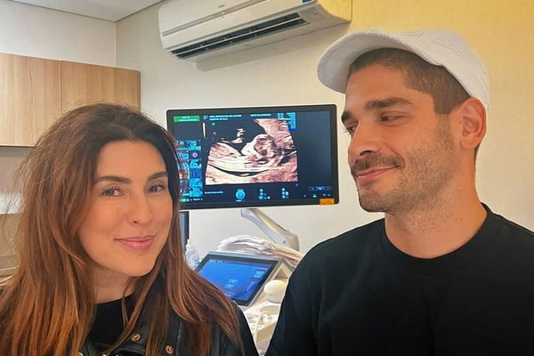 Foto colorida de Fernanda Paes Leme e Victor Sampaio se entreolhando com um ultrassom na tela ao fundo no meio dos dois - Metrópoles