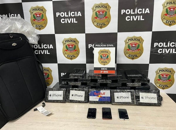 Foto colorida mostra tabletes de droga embalados em sacos pretos e celulares sobre mesa. Ao fundo, está um banner da Polícia Civil