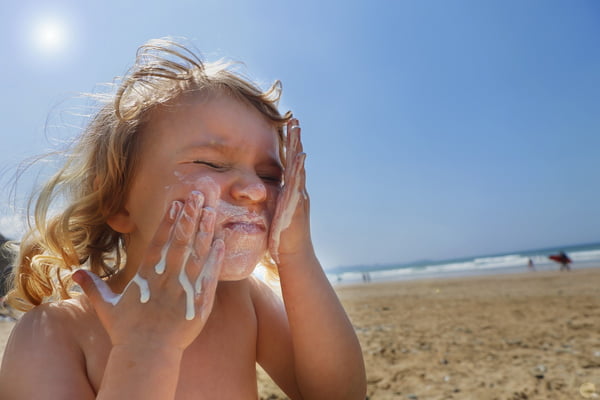 Fotografia mostra criança loira na praia passando muito filtro solar no rosto - Metrópoles