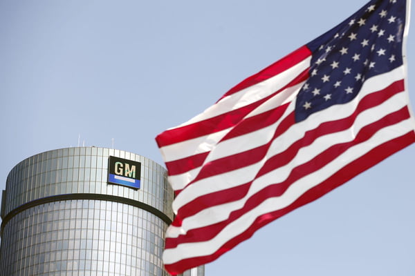 Imagem colorida da bandeira dos Estados Unidos. Ao fundo, fábrica da General Motors - Metrópoles