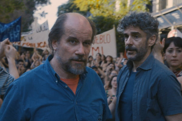 Foto colorida dos atores Marcelo Subiotto e Leonardo Sabaraglia em cena de Puan. Ao fundo, milhares de pessoas empunhão cartazes - Metrópoles