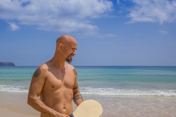 Foto colorida mostra homem calvo, careca em praia com sol intenso