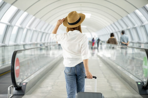 Imagem de mulher de chapéu carregando mala de rodinha em aeroporto - Metrópoles