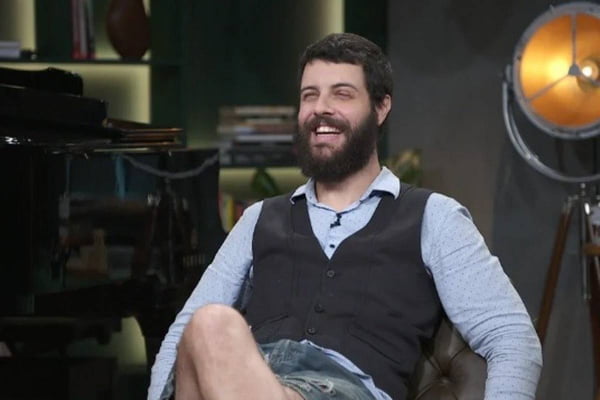 Foto colorida de Diogo Defante sentado e sorrindo, com colete e blusa de manga - Metrópoles