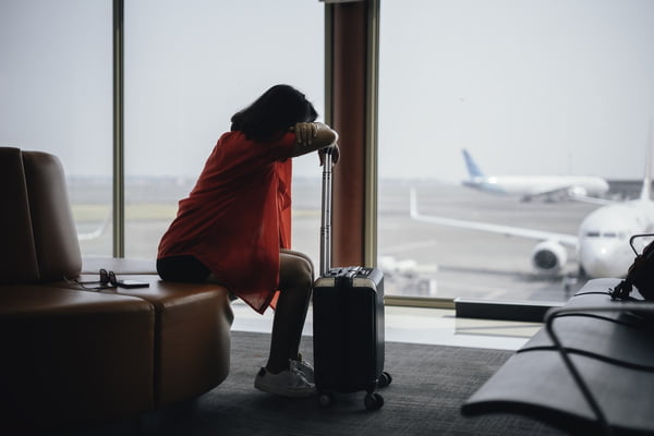 Imagem colorida de uma mulher de blusa vermelha apoiando-se em uma mala de viagem, em um aeroporto - Metrópoles